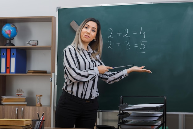 joven profesora de pie cerca de la pizarra en el aula explicando la lección sosteniendo el puntero presentando con el brazo de su mano sonriendo confiada