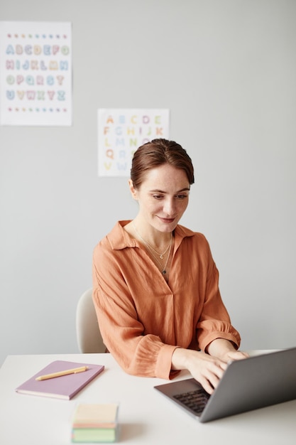 Joven profesora sentada en la mesa y escribiendo en la computadora portátil que se prepara para la lección