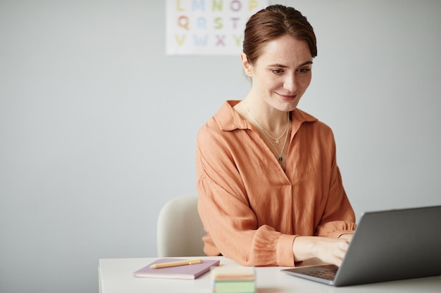 Joven profesora sentada en su lugar de trabajo y trabajando en línea en una computadora portátil que se prepara para las lecciones en la escuela