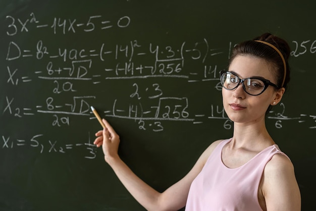 Joven profesora seria explica fórmulas matemáticas en la universidad al lado de la pizarra