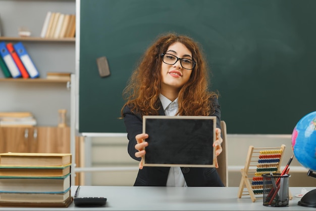 una joven profesora sonriente con gafas sosteniendo una mini pizarra a la cámara sentada en un escritorio con herramientas escolares en el aula