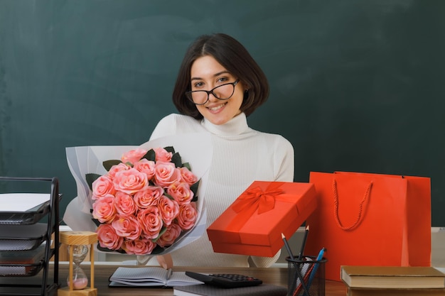 una joven profesora sonriente sosteniendo un ramo con una caja de regalo sentada en el escritorio con herramientas escolares en el aula