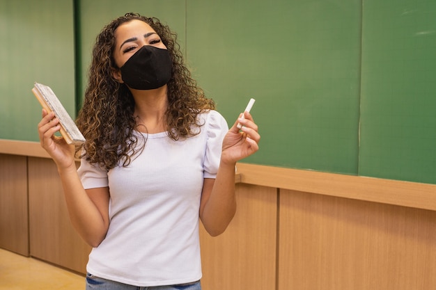 Joven profesora con tiza y goma de borrar en la mano con mascarilla quirúrgica en nueva normalidad.