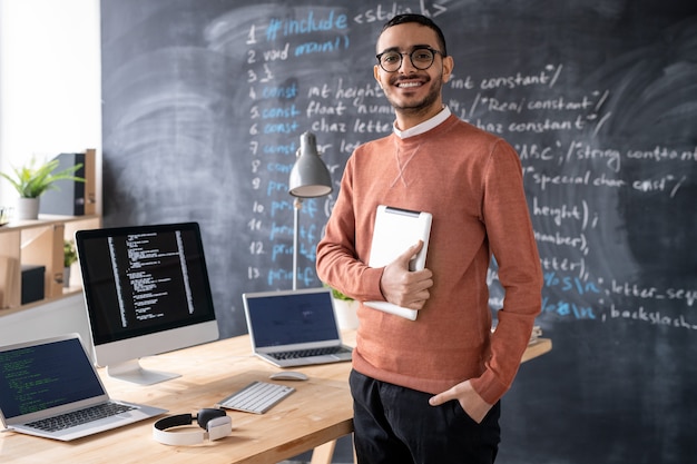 Joven programador alegre con dispositivo móvil de pie junto a la mesa con ordenadores portátiles y computadora con pizarra