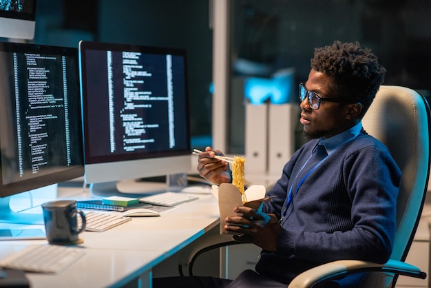 Joven programador hambriento comiendo wok chino frente a la pantalla de la computadora con datos codificados