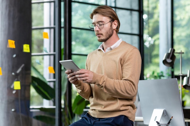 Joven programador de pensamiento exitoso dentro de la oficina de estilo loft usa una computadora de tableta joven rubio