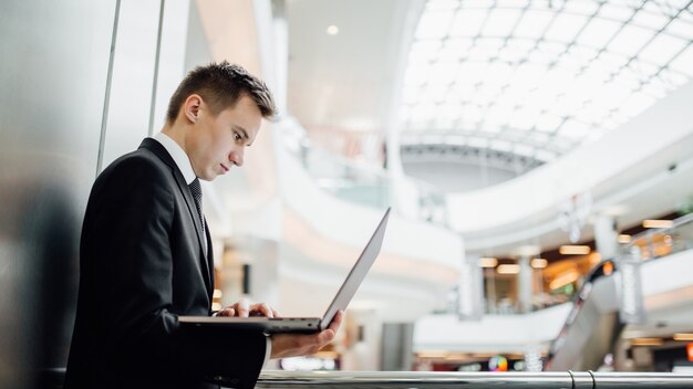 Joven programador, trabajando en una computadora portátil, en el centro de negocios, interior, vista de perfil, vestido con traje negro en el centro comercial,