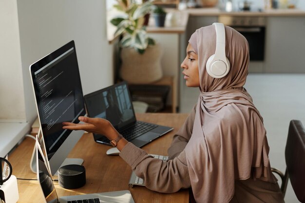 Joven programadora musulmana señalando datos en la pantalla de la computadora mientras está sentada en el lugar de trabajo