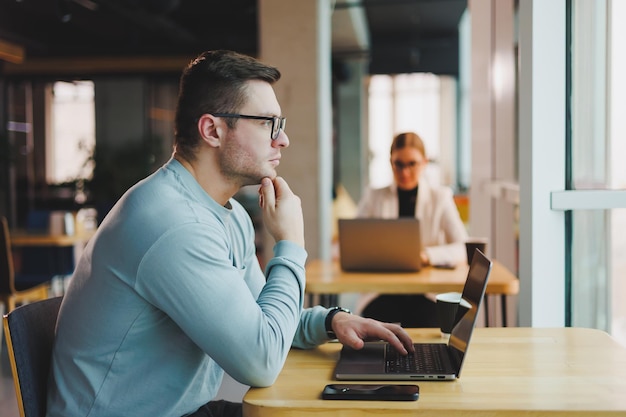 Un joven prometedor con ropa informal se sienta en una mesa y trabaja en una computadora portátil Espacio de trabajo para trabajo remoto