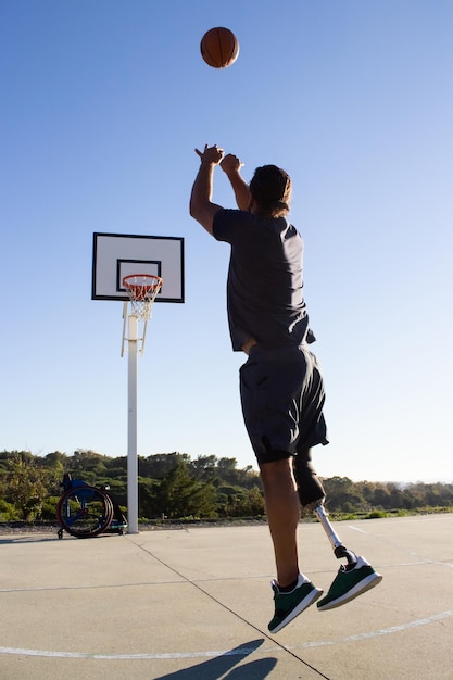 Joven con prótesis de pierna lanzando baloncesto al aro. Vista posterior del jugador de baloncesto masculino con discapacidad saltando mientras juega afuera. Silla de ruedas en segundo plano. Concepto deportivo para amputados