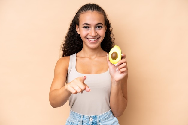 Una joven que sostiene un aguacate aislado de fondo beige te señala con una expresión de confianza