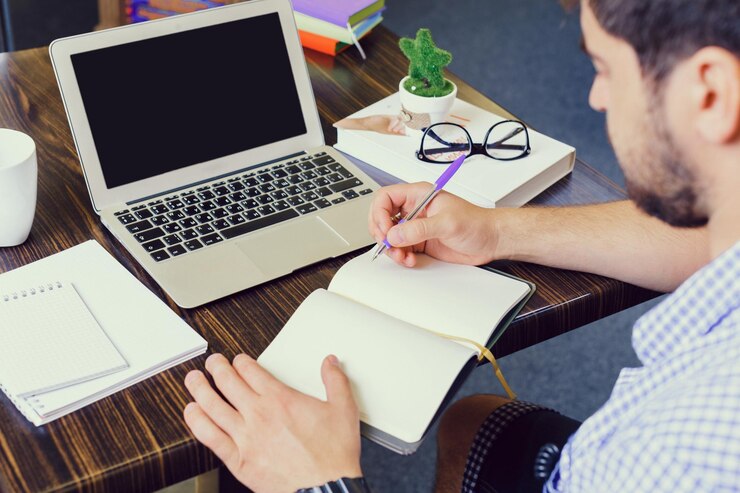 Foto un joven que trabaja en un escritorio escribiendo notas en un cuaderno mientras usa una computadora portátil en un entorno de estudio