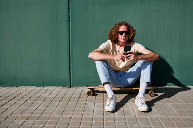 un joven que usa su teléfono inteligente y está sentado en su patineta o longboard con una pared detrás de él