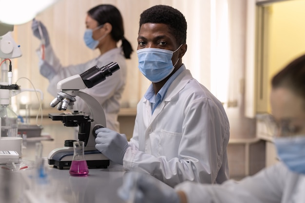 Joven químico africano masculino en workear protectora usando microscopio