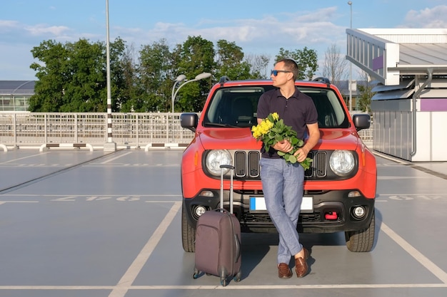 Un joven con un ramo de flores y una maleta se para afuera de su auto al aire libre