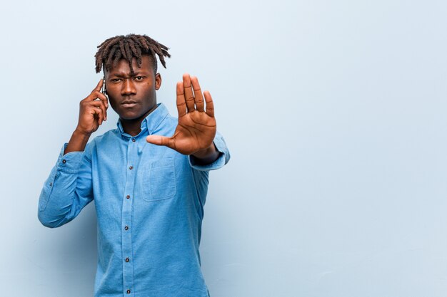 Joven rasta negro sosteniendo un teléfono de pie con la mano extendida que muestra la señal de stop, impidiéndole.