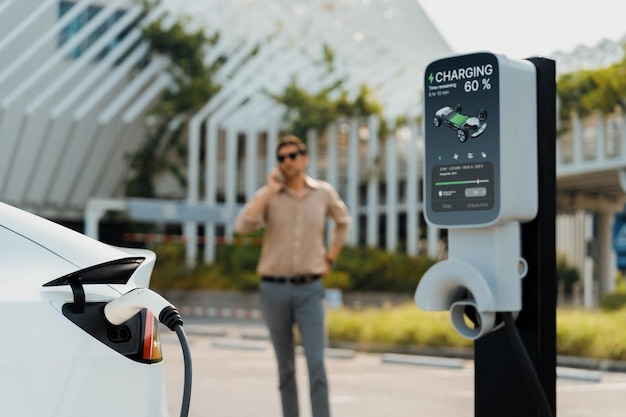 Un joven recarga un vehículo eléctrico EV en el estacionamiento del centro comercial de la ciudad verde mientras habla por teléfono Estilo de vida urbano sostenible para un vehículo EV ecológico con estación de carga de batería
