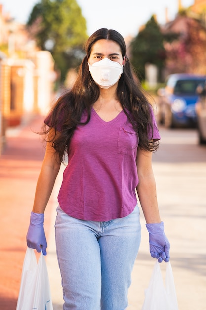 Joven regresa de compras con bolsas en las manos con guantes médicos y máscara de virus durante la pandemia de coronavirus en la calle