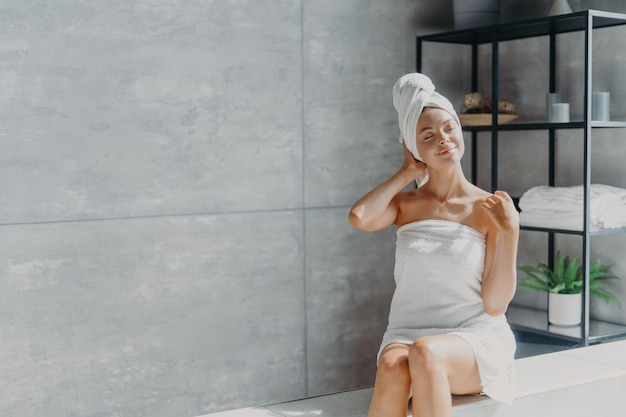 Una joven y relajada modelo caucásica usa una toalla envuelta en la cabeza y se siente refrescada después de tomar una ducha, tiene poses saludables de piel limpia y suave en un baño acogedor Concepto de belleza e higiene para mujeres