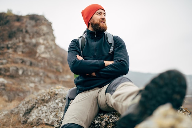 Joven relajándose en las montañas con mochila de viaje y con sombrero rojo El viajero barbudo tiene un descanso después de hacer senderismo durante las vacaciones mira hacia otro lado Estilo de vida de la gente de viaje