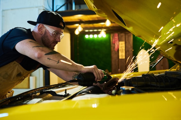 Joven reparador agachado sobre el motor del automóvil mientras usa una amoladora eléctrica