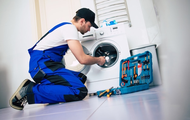 El joven reparador guapo en traje de trabajador con la caja de herramientas profesionales está arreglando la lavadora en el baño