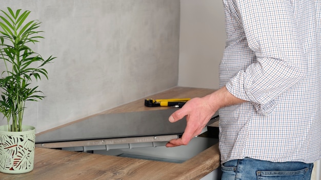 Un joven reparador instala una placa de inducción negra en una moderna cocina blanca de estilo escandinavo con una pared de hormigón. Hombre electricista, hágalo usted mismo. Tareas del hogar.