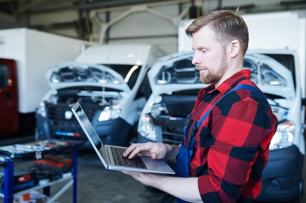 Joven reparador profesional del centro de servicio de automóviles usando una computadora portátil mientras navega en el sitio web o realiza pedidos en línea