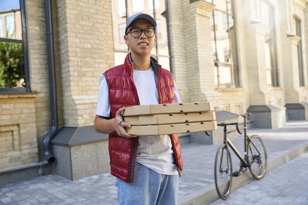 Joven repartidor asiático alegre sosteniendo cajas con pizza y l