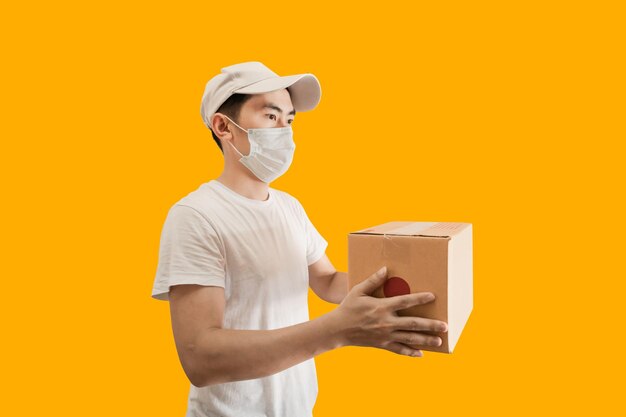 Joven repartidor asiático con gorra y camiseta blanca en blanco sosteniendo una caja postal de paquetes aislada en un fondo amarillo concepto de servicio de entrega expreso