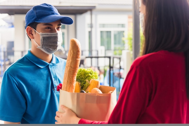 Un joven repartidor asiático con uniforme usa una máscara protectora para la cara que hace el servicio de comestibles dando comida fresca a una cliente que recibe la casa bajo el coronavirus pandémico, Volver al nuevo concepto normal