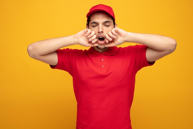 joven repartidor cansado con uniforme y gorra manteniendo las manos cerca de la boca bostezando con los ojos cerrados y estirándose aislado sobre fondo amarillo