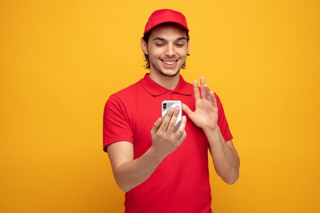 un joven repartidor sonriente con uniforme y gorra sosteniendo un teléfono móvil hablando a través de una videollamada mostrando un gesto de saludo aislado en un fondo amarillo