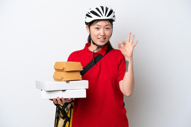 Joven repartidora china tomando comida para llevar aislada de fondo blanco mostrando el signo de ok con los dedos