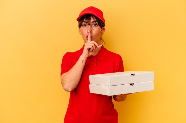 Joven repartidora sosteniendo pizzas aisladas de fondo amarillo guardando un secreto o pidiendo silencio.