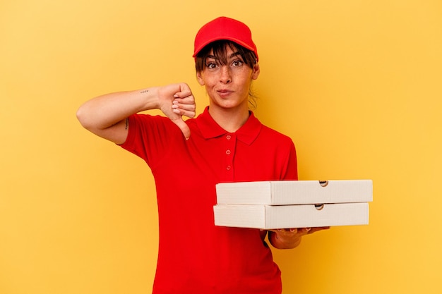 Joven repartidora sosteniendo pizzas aisladas de fondo amarillo mostrando un gesto de aversión, pulgares hacia abajo. Concepto de desacuerdo.