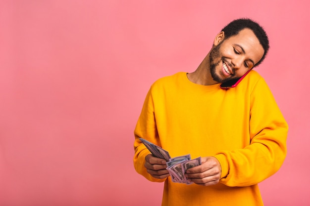 Joven rico en casual sosteniendo dinero billetes de un dólar y teléfono móvil con sorpresa aislado sobre pared rosa