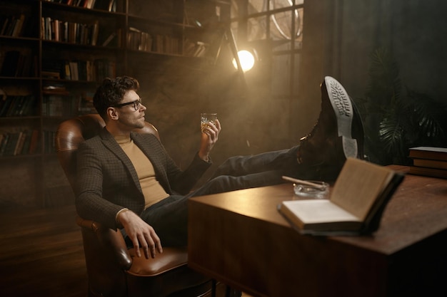 Joven rico con traje elegante bebiendo whisky descansando en un sillón de lujo poniendo un pie en la mesa en el gabinete de casa