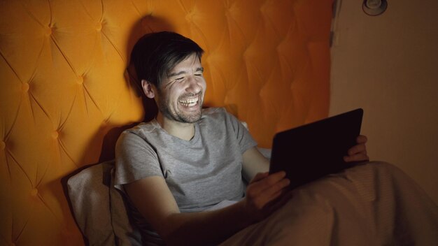 Joven riendo usando una tableta navegando en las redes sociales acostado en la cama en casa antes de dormir