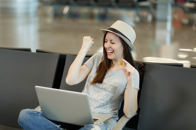 Joven riendo viajero turista mujer con sombrero sentado trabajando en un portátil sosteniendo bitcoin haciendo gesto de ganador esperando en el vestíbulo del aeropuerto