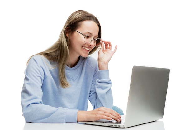 Una joven risa con gafas ve en una mesa con un portátil y mira el monitor. Formación, trabajo a distancia y blogs. Aislado en una pared blanca. Espacio para texto.