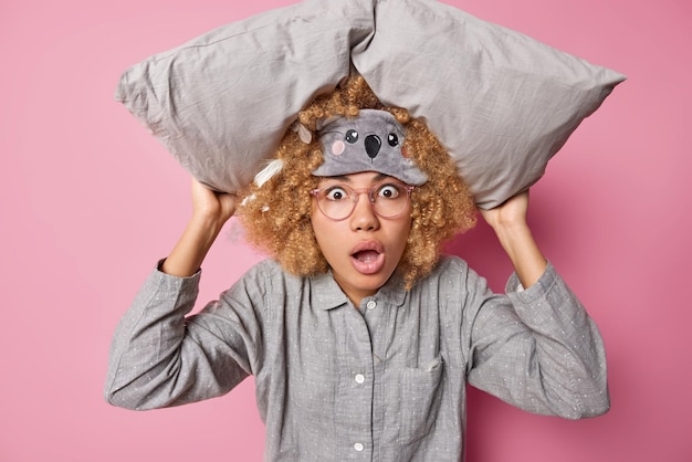 Foto una joven rizada sorprendida se despierta durante la noche después de ver que la pesadilla sostiene una almohada sobre la cabeza y mira asombrada con una expresión asustada que usa una venda en los ojos y un pijama aislado sobre un fondo rosado