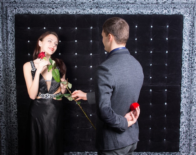 Joven romántico pidiendo a una hermosa joven con un elegante vestido de cóctel negro que se case con él presentándole una rosa antes de producir el anillo.