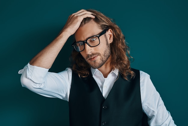 Joven romántico en ropa elegante y gafas mirando a la cámara y manteniendo la mano en el cabello mientras está de pie contra la pared verde