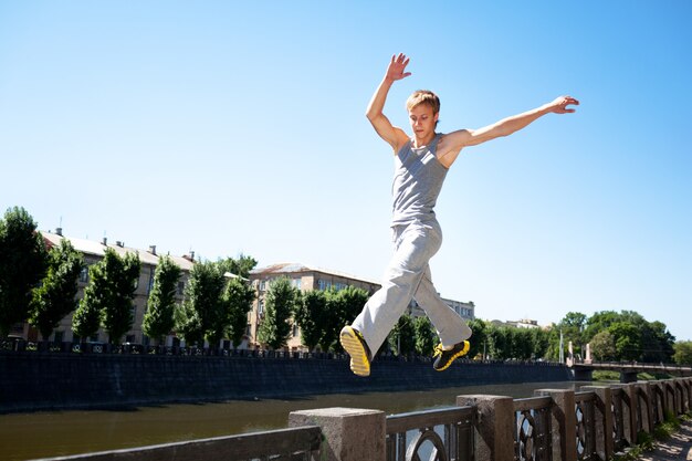 Joven en ropa deportiva saltando y practicando parkour afuera en la valla de piedra sobre el terraplén del río en un claro día de verano