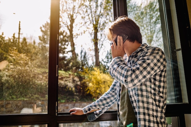 Joven con ropa informal hablando por un teléfono móvil por la mañana en una ventana con espacio para copiar