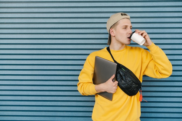 un joven con ropa informal y una laptop en las manos se para en un fondo azul tomando café