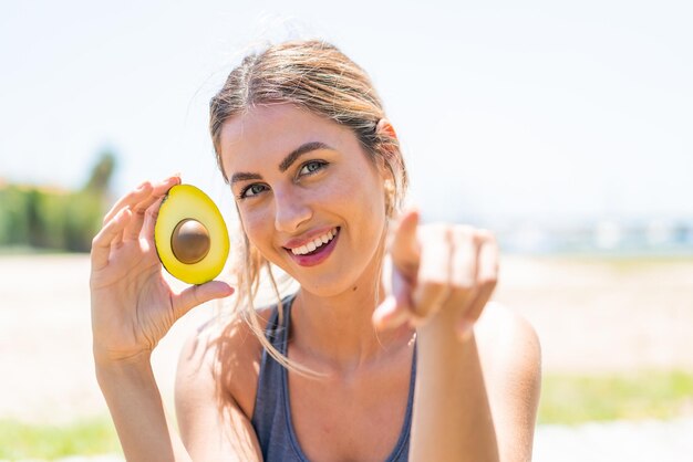 Foto una joven rubia con un aguacate al aire libre te señala con un dedo con una expresión de confianza