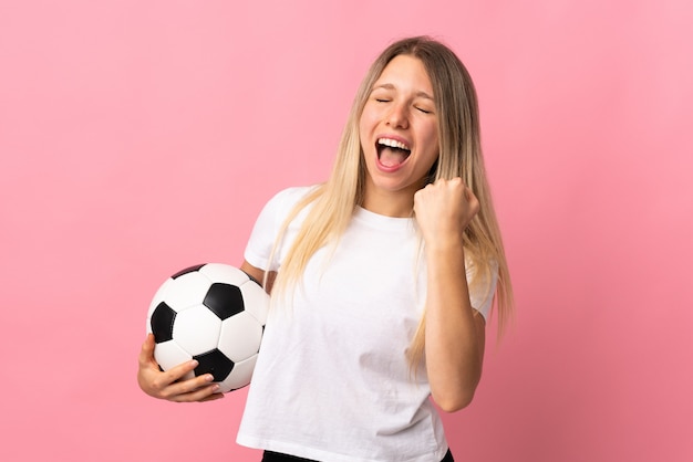 Joven rubia aislada en la pared rosa con balón de fútbol celebrando una victoria