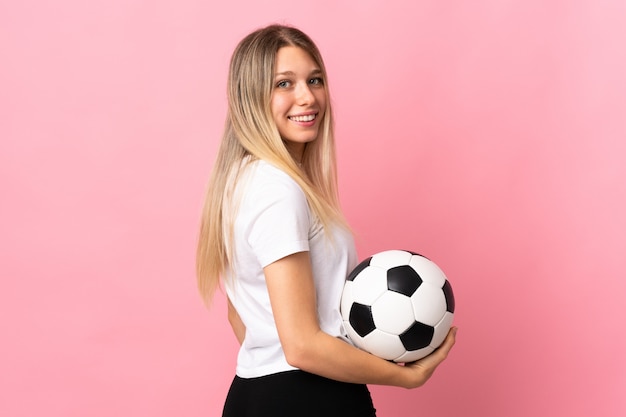 Joven rubia aislada en pared rosa con balón de fútbol